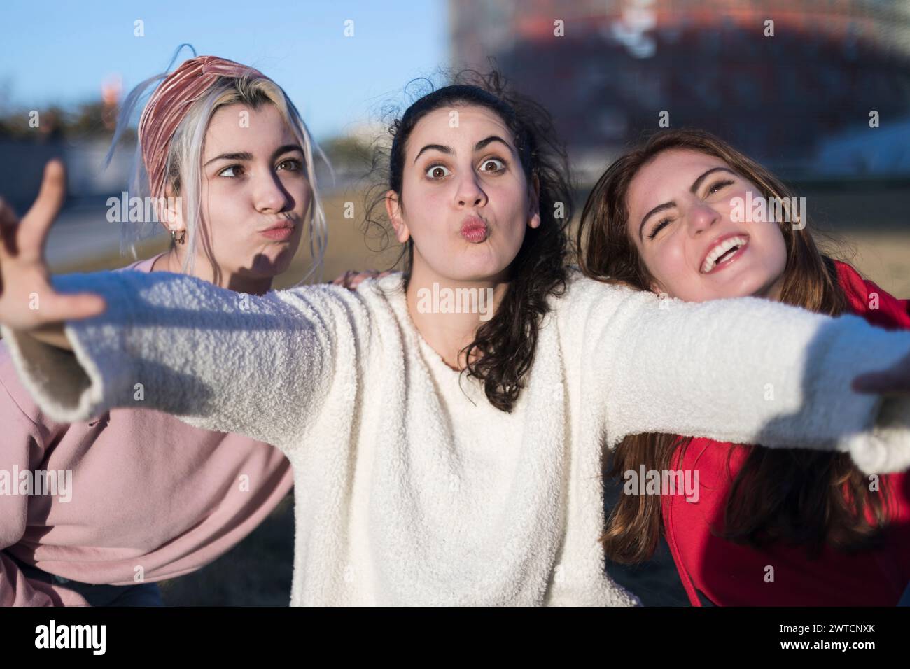 Three young women smile outdoors, arms extended in a carefree pose. Middle with pink sweater, left in red, right in white. Sunny urban backdrop. Stock Photo