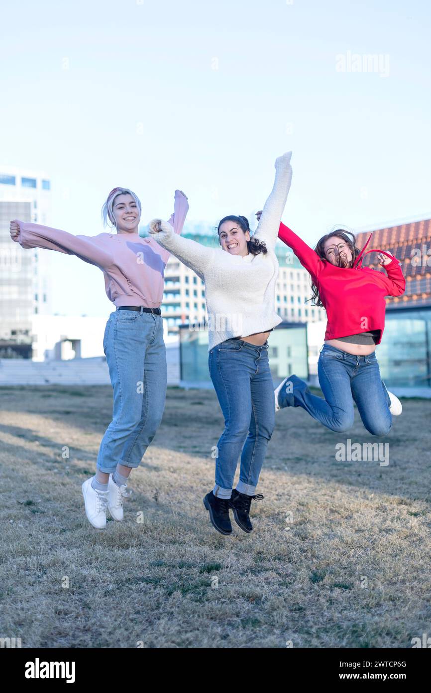 Three individuals woman, dressed in jeans and sweaters, are captured mid-jump in an outdoor field. Arms extended, they express freedom and joy. Urban Stock Photo
