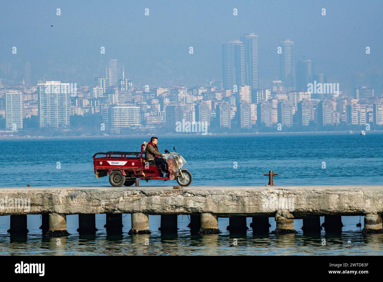 A lman on a delivery truck drives along a jetty with a backdrop of the skyscrapers and office buildings of modern Istanbul, Turkey, in the distance. Stock Photo