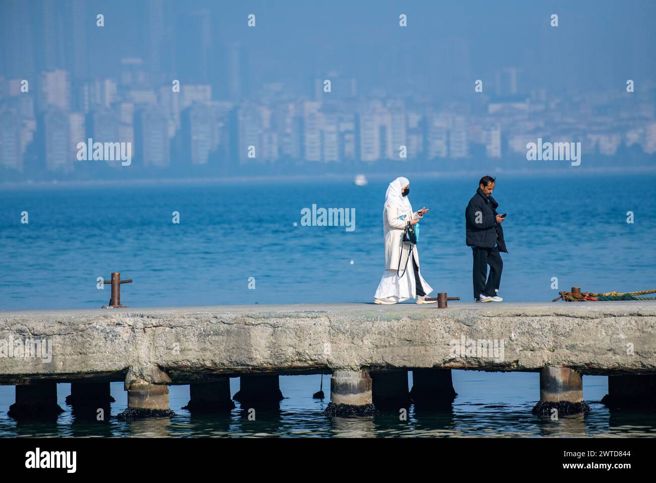 A lady and a man walk along a jetty with a backdrop of the skyscrapers and office buildings of modern Istanbul, Turkey, in the distance. Stock Photo