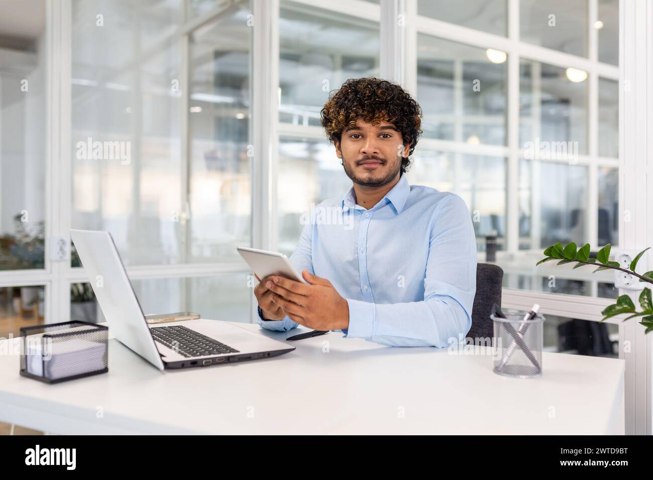A focused young man in a blue shirt engaged in multitasking with a laptop and tablet at a bright office desk. Stock Photo