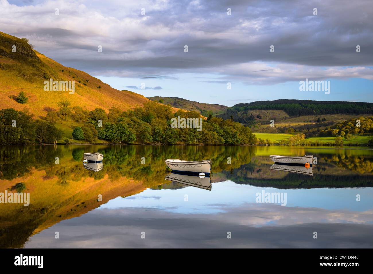 Three wooden rowing boats sit motionless upon the mirror like waters of Ullswater on a calm spring morning. Stock Photo