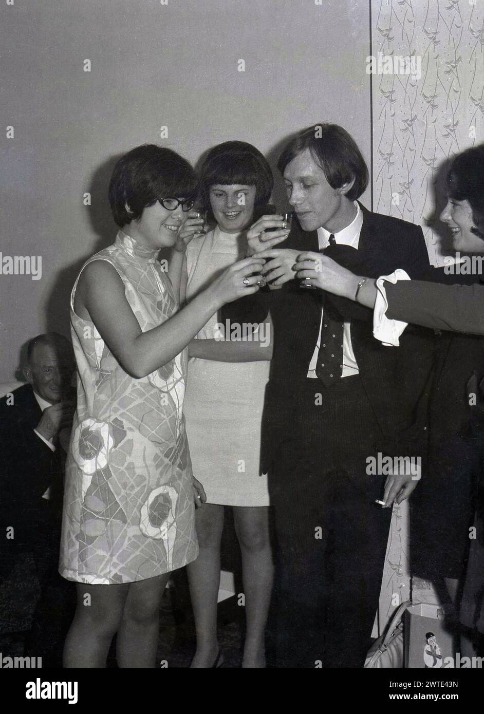 1960s, historical, 21st birthday celebrations, cheers... clinking glasses, friends of the birthday girl, in the sleeves patterned dress, toast her health and happiness on her special day, England, UK. Stock Photo