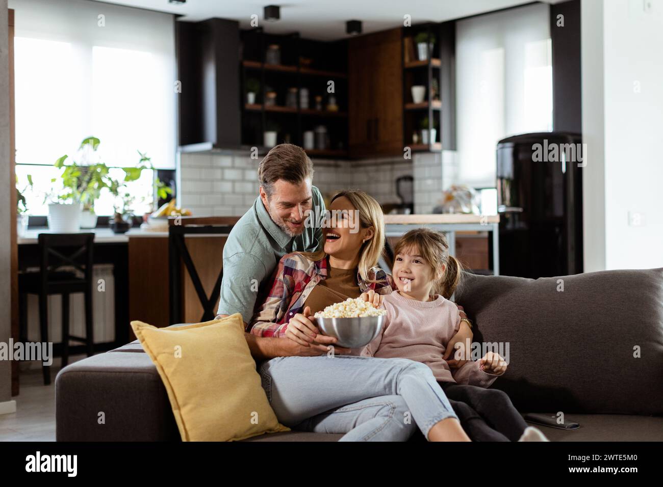 A family of three is comfortably nestled on a couch, their faces reflecting excitement and attentiveness as they share a bowl of popcorn during a susp Stock Photo