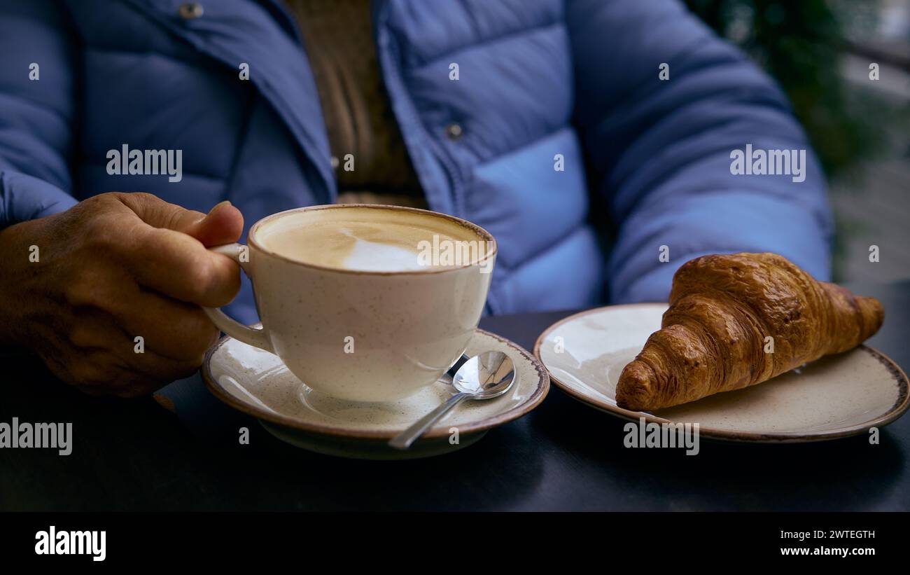 Woman holding a cup of cappuccino and a croissant in a plate nearby on the table Stock Photo