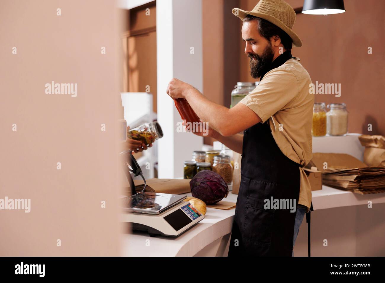 Caucasian male shopkeeper, who wears a stylish hat and black apron, stands behind the checkout counter holding a jar filled with pasta sauce. Vendor analyzing glass container at cashier desk. Stock Photo