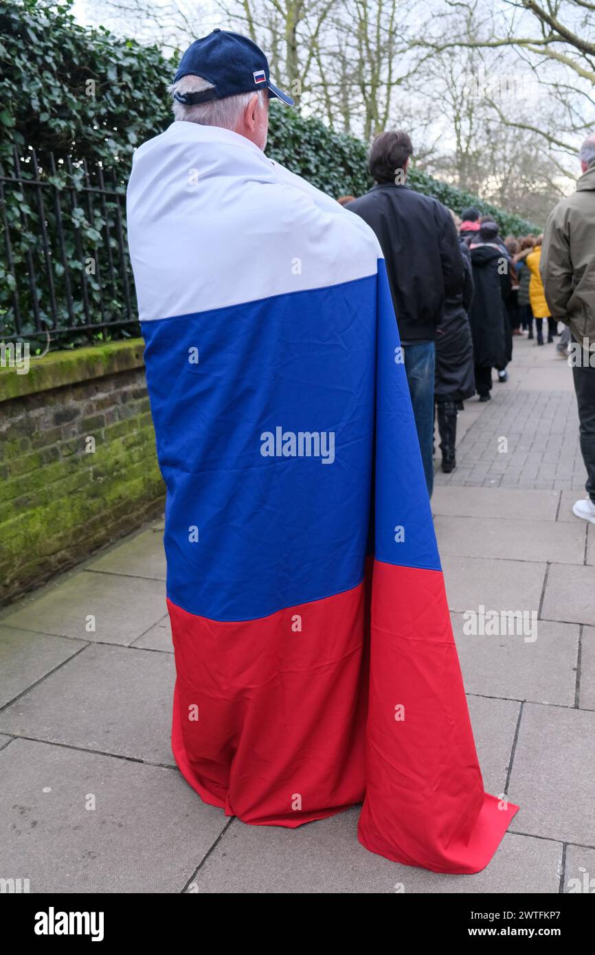 London, UK. 17th March, 2024. A Putin supporter draped in a large Russian flag waits in the voters' queue with his wife. Thousands of voters descended on the Russian Embassy in Kensington with some voters queuing for four hours before they cast their ballot. Many waiting were seen draped in the pro-democracy blue and white flag as Yulia, the wife of the late opposition leader Alexei Navalny, urged voters to turn up in a 'Noon against Putin' protest, whilst Navalny's team asked for individuals to spoil their ballot papers. Credit: Eleventh Hour Photography/Alamy Live News Stock Photo