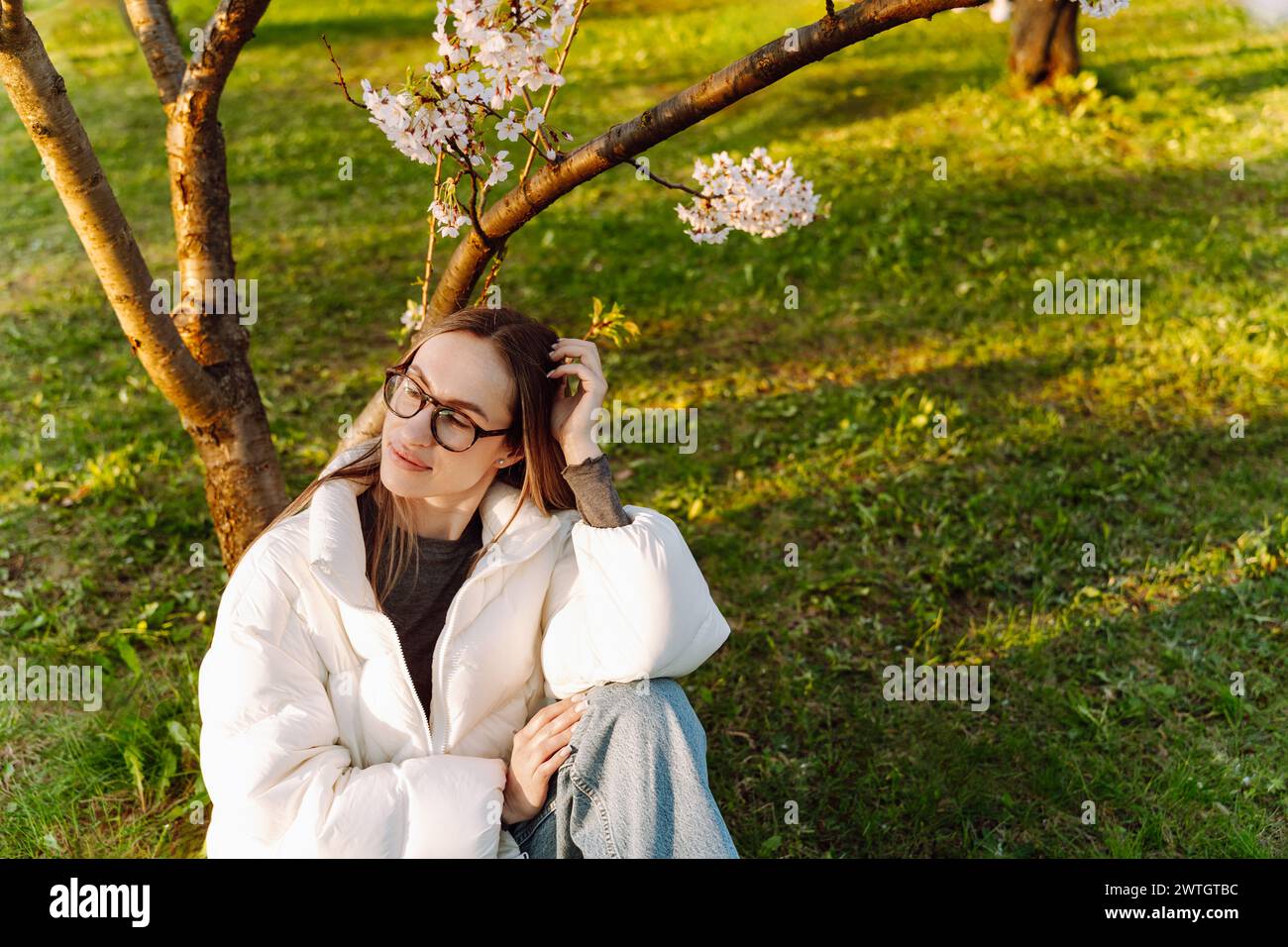 Beautiful young woman in glasses and white coat sits near blooming tree Stock Photo