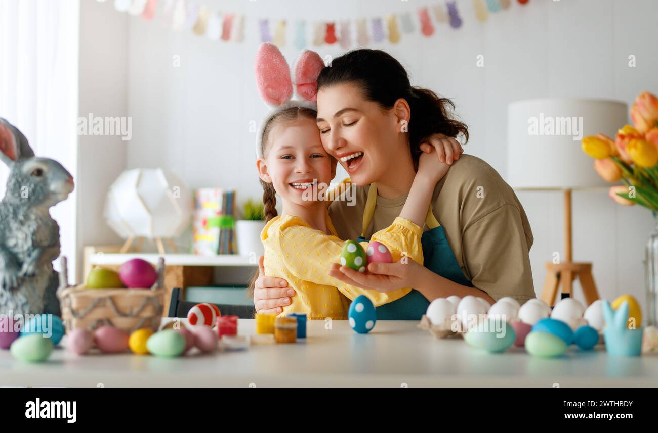 Mother and her daughter painting eggs. Happy family preparing for Easter. Cute little child girl wearing bunny ears. Stock Photo