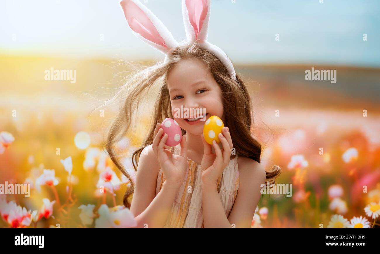 Beautiful child with painting eggs outdoors. Happy family celebrating Easter in nature. Cute little girl is wearing bunny ears. Stock Photo