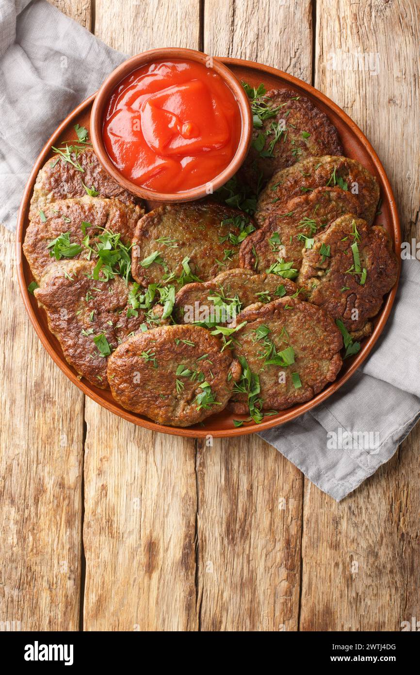 Fried beef liver patties served with tomato sauce and herbs close-up in a plate on the table. Vertical top view from above Stock Photo