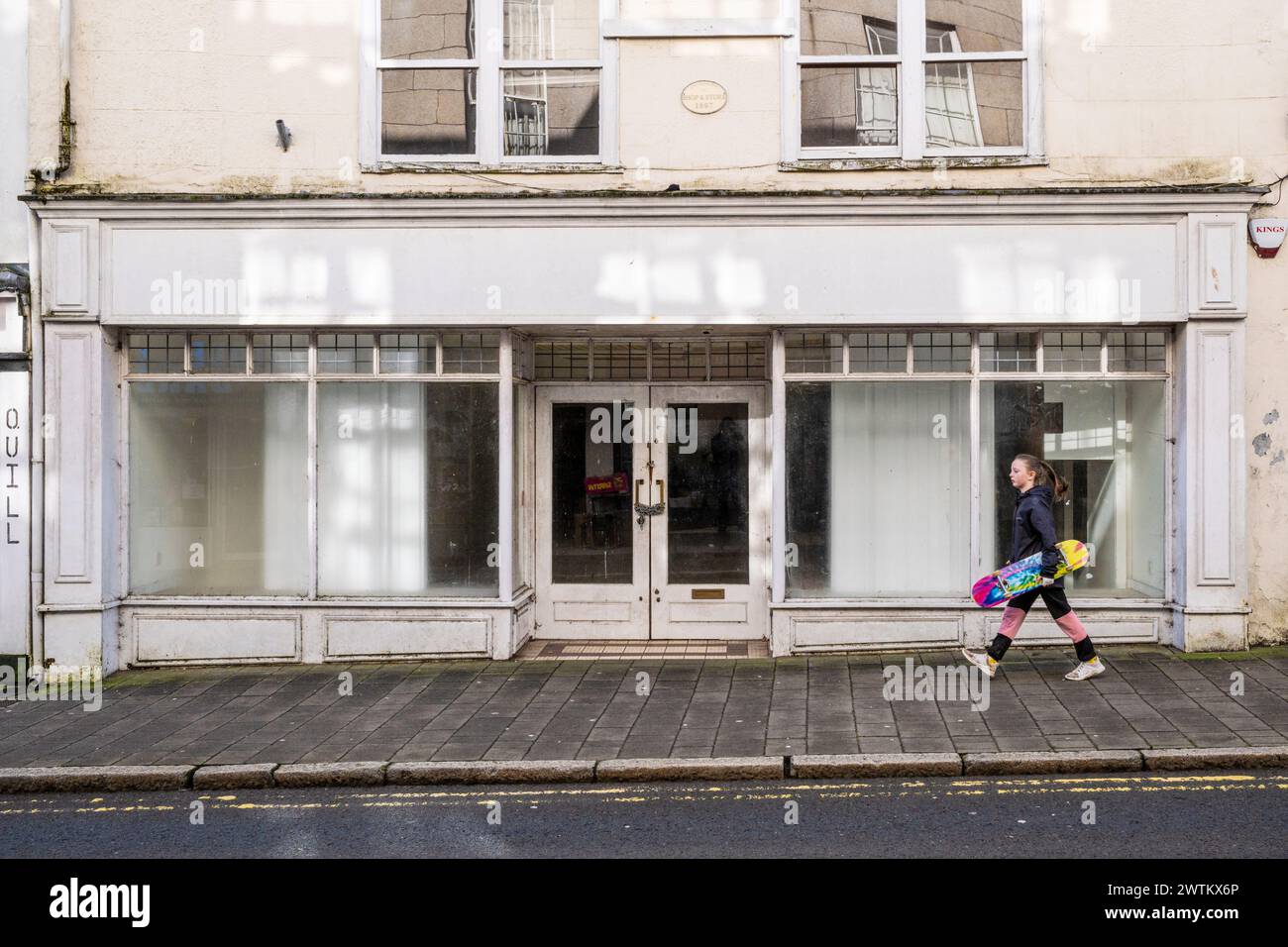 A young girl carrying a colourful colorful skateboard and walking past an empty closed shop store in Fore street in Bodmin Town centre in Cornwall in Stock Photo