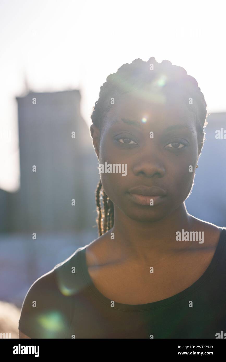 An African woman stands backlit by the setting sun, her silhouette partially outlined against the soft glow of the evening sky. The suns rays filter through her hair, creating a natural halo effect around her head. Her expression is solemn and introspective, with the city skyline forming a muted backdrop. The warm backlighting and the slight lens flare add a dreamy and reflective quality to the image. Backlit African Woman with Urban Silhouette. High quality photo Stock Photo