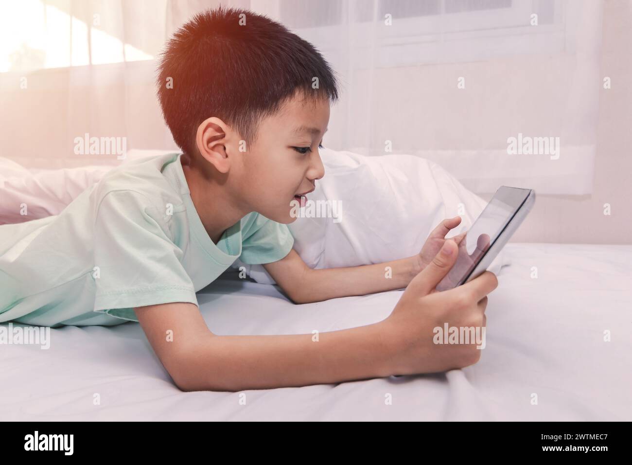 little boy looking at a tablet and playing games while lying on the bed ...