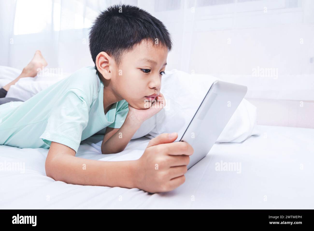 little boy looking at a tablet and playing games while lying on the bed ...