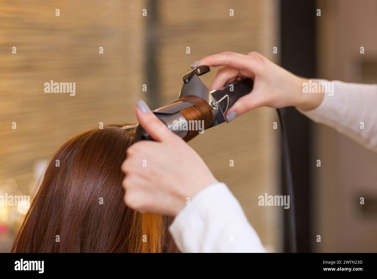Hairdresser makes hairstyle girl with long red hair in a beauty salon. Create curls with curling irons. Professional hair care. Stock Photo