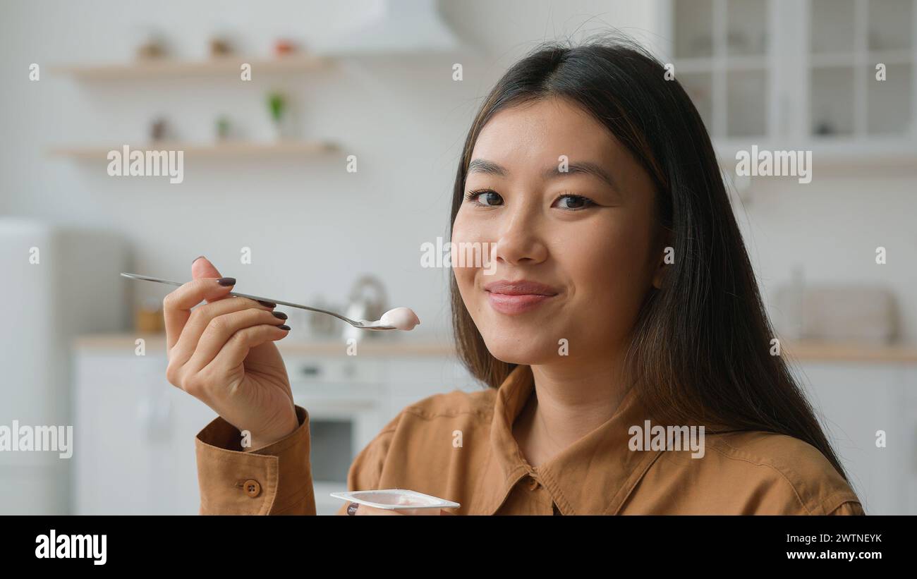 Asian korean woman girl tasting healthy nutrition eat creamy fruit yogurt in kitchen chinese girl smiling at camera eating dairy lactose dessert with Stock Photo