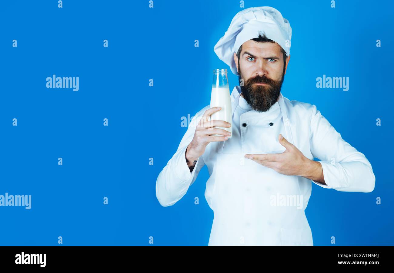 Professional male chef or confectioner pointing hand at fresh milk for making desserts. Serious bearded cook in chef hat and uniform with bottle of Stock Photo