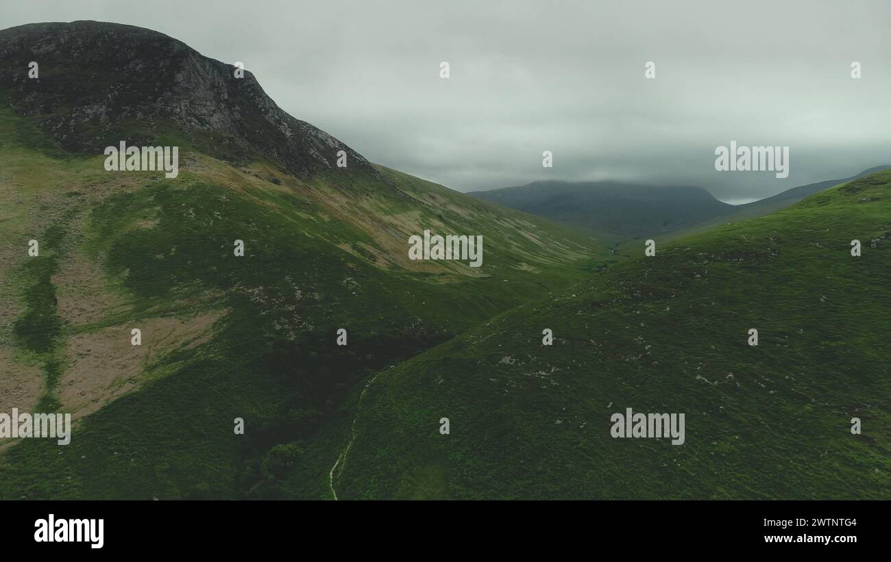 Scotland mountain green ranges aerial view. Path between greenery grassy hills. Grey clouds wrapped top of mounts in summer day. Wild untouched nature scenery of island Arran. Footage Shot in 2K (QHD) Stock Photo