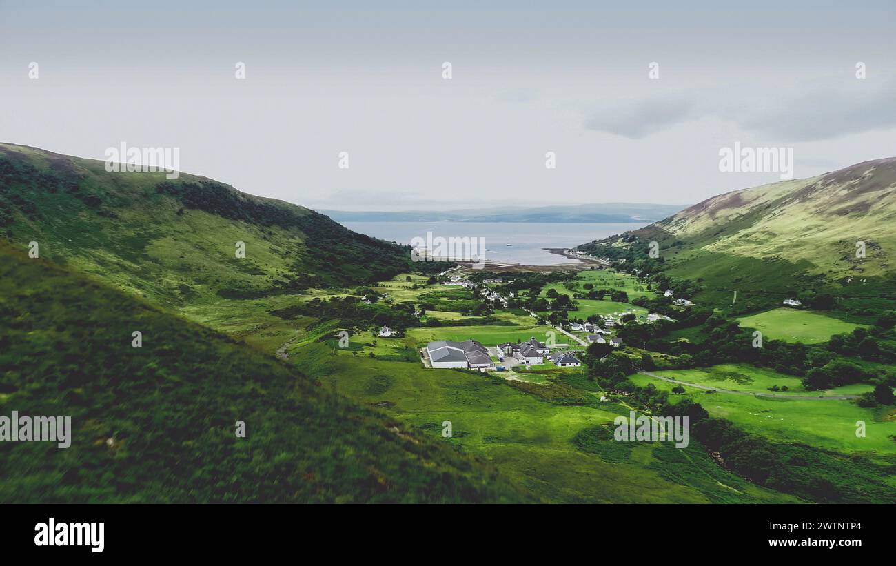 Scotland village aerial shot: mountain, greenery valley. Scottish hillside town with sea lake, castle ruins. Loch Ranza resort and distillery in Arran Island, UK. Footage shooting Stock Photo