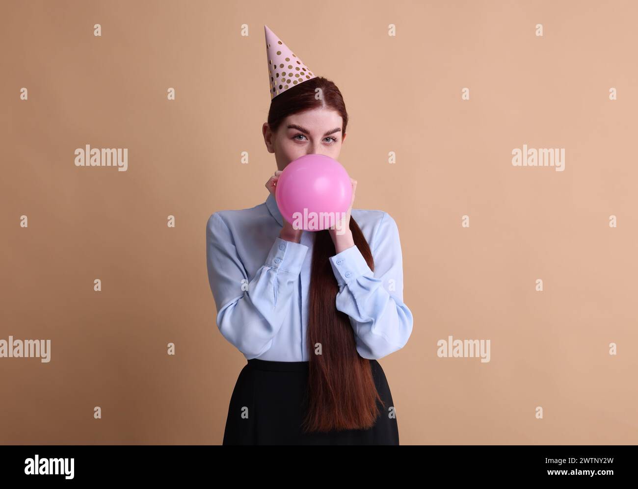 Young woman in party hat blowing balloon on beige background Stock Photo