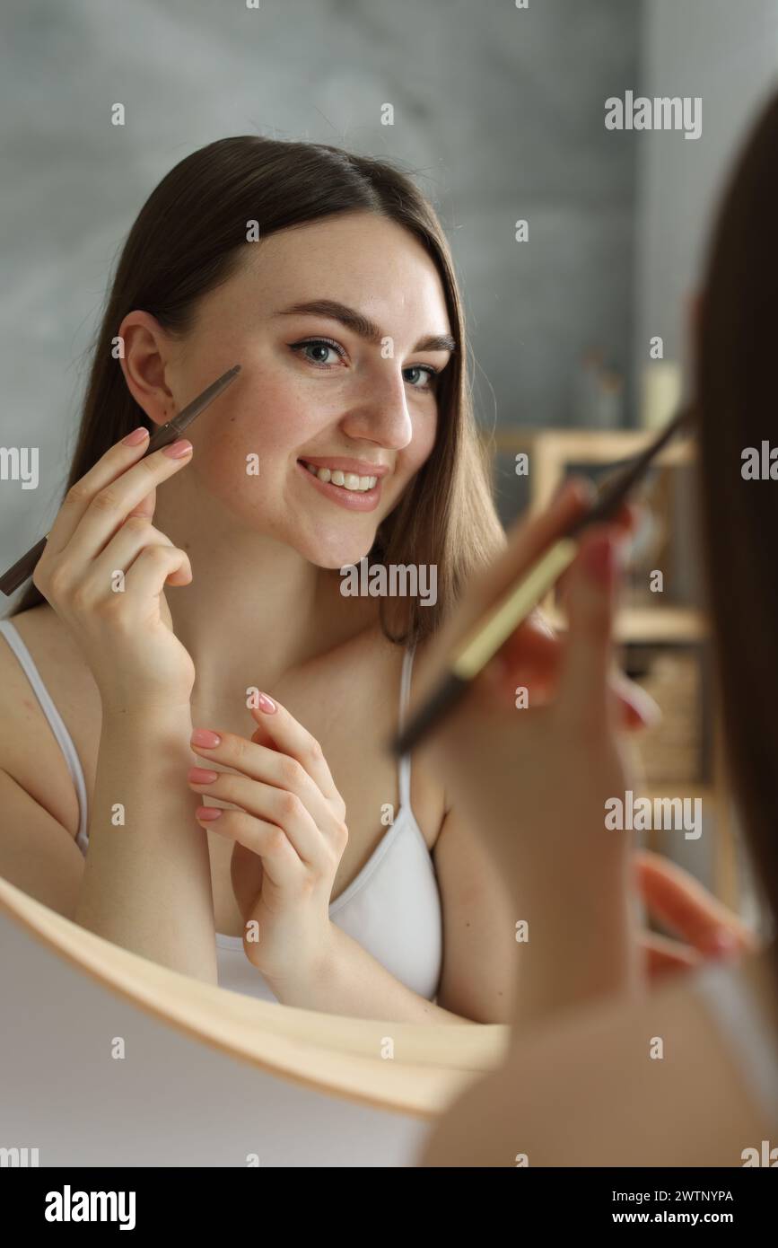 Smiling woman drawing freckles with pen near mirror indoors Stock Photo