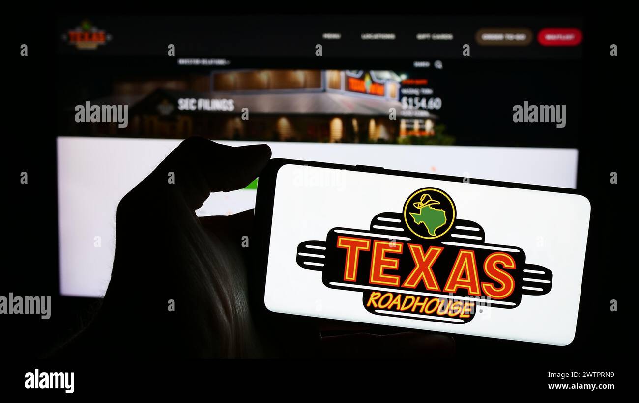 Person holding smartphone with logo of US steakhouse restaurant company Texas Roadhouse Inc. in front of website. Focus on phone display. Stock Photo