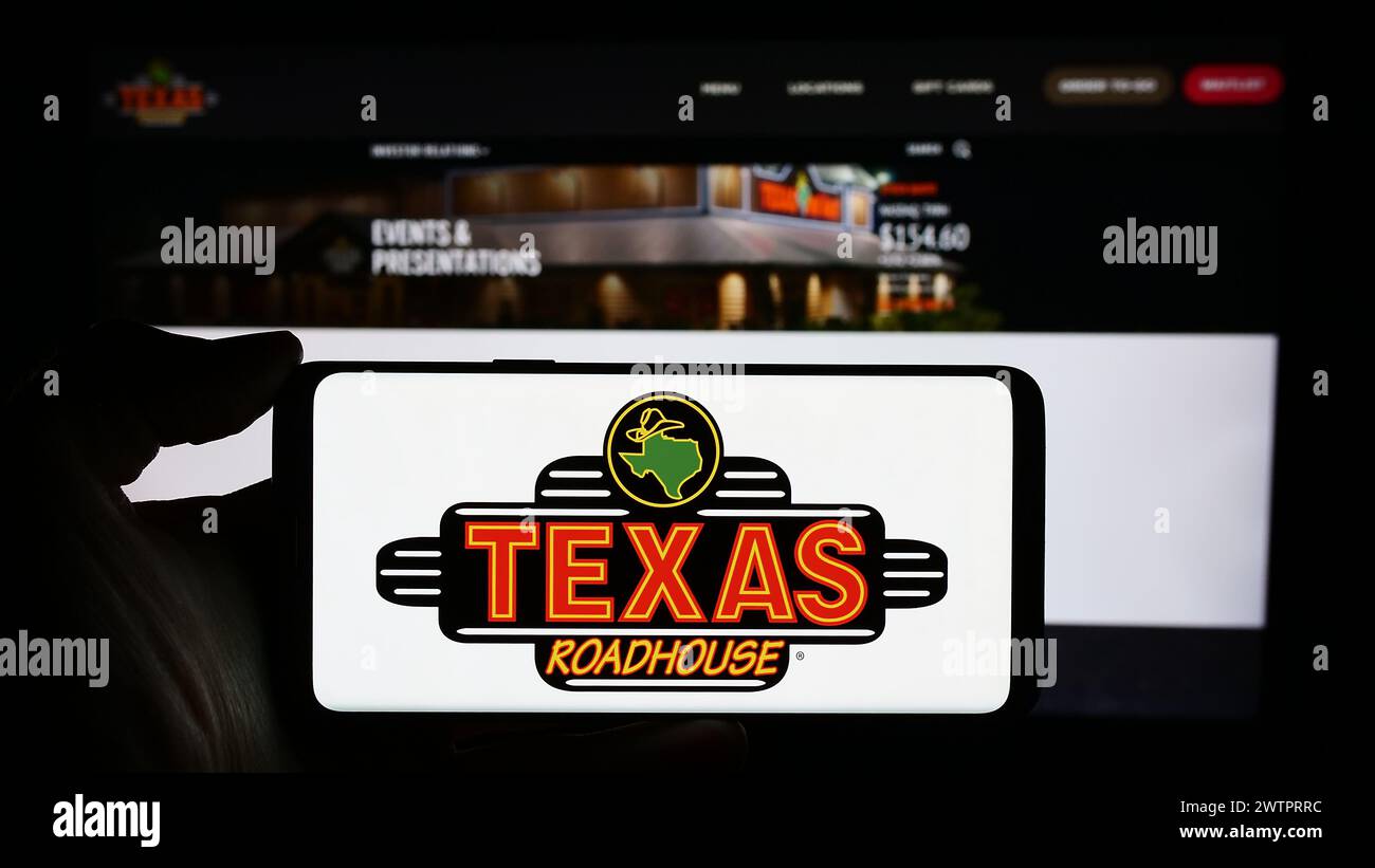 Person holding cellphone with logo of US steakhouse restaurant company Texas Roadhouse Inc. in front of business webpage. Focus on phone display. Stock Photo