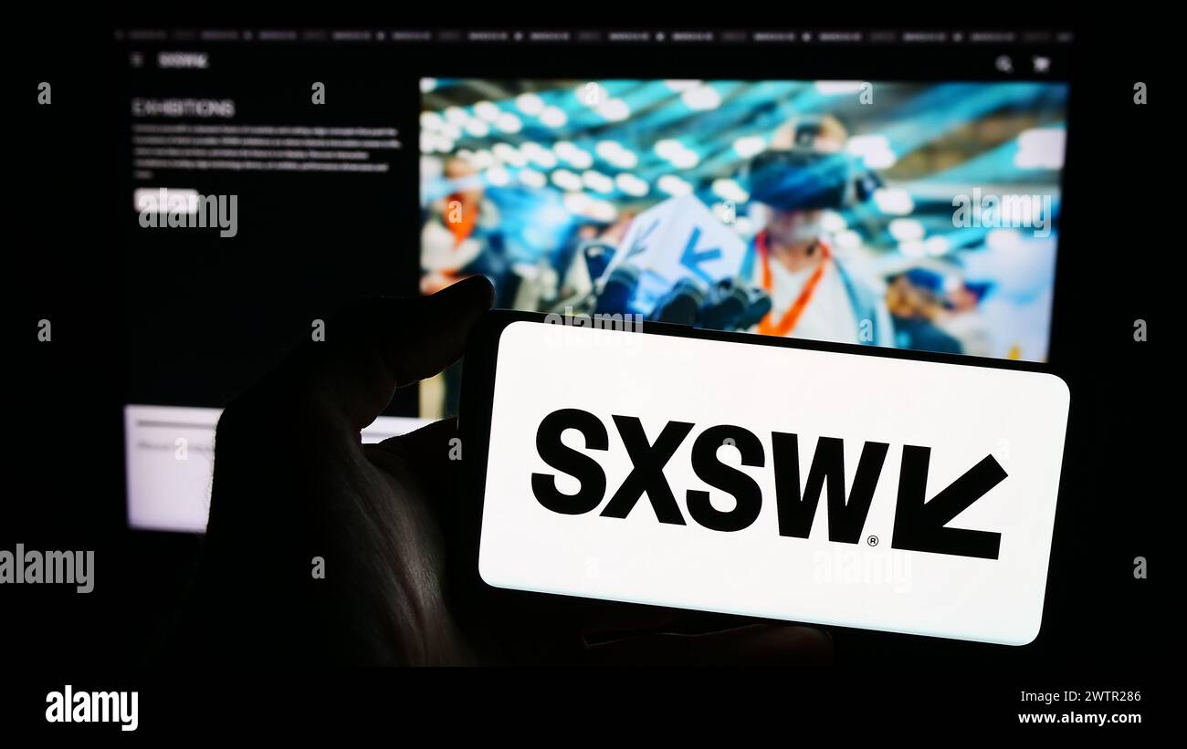 Person holding smartphone with logo of annual film and media event South by Southwest (SXSW) in front of website. Focus on phone display. Stock Photo