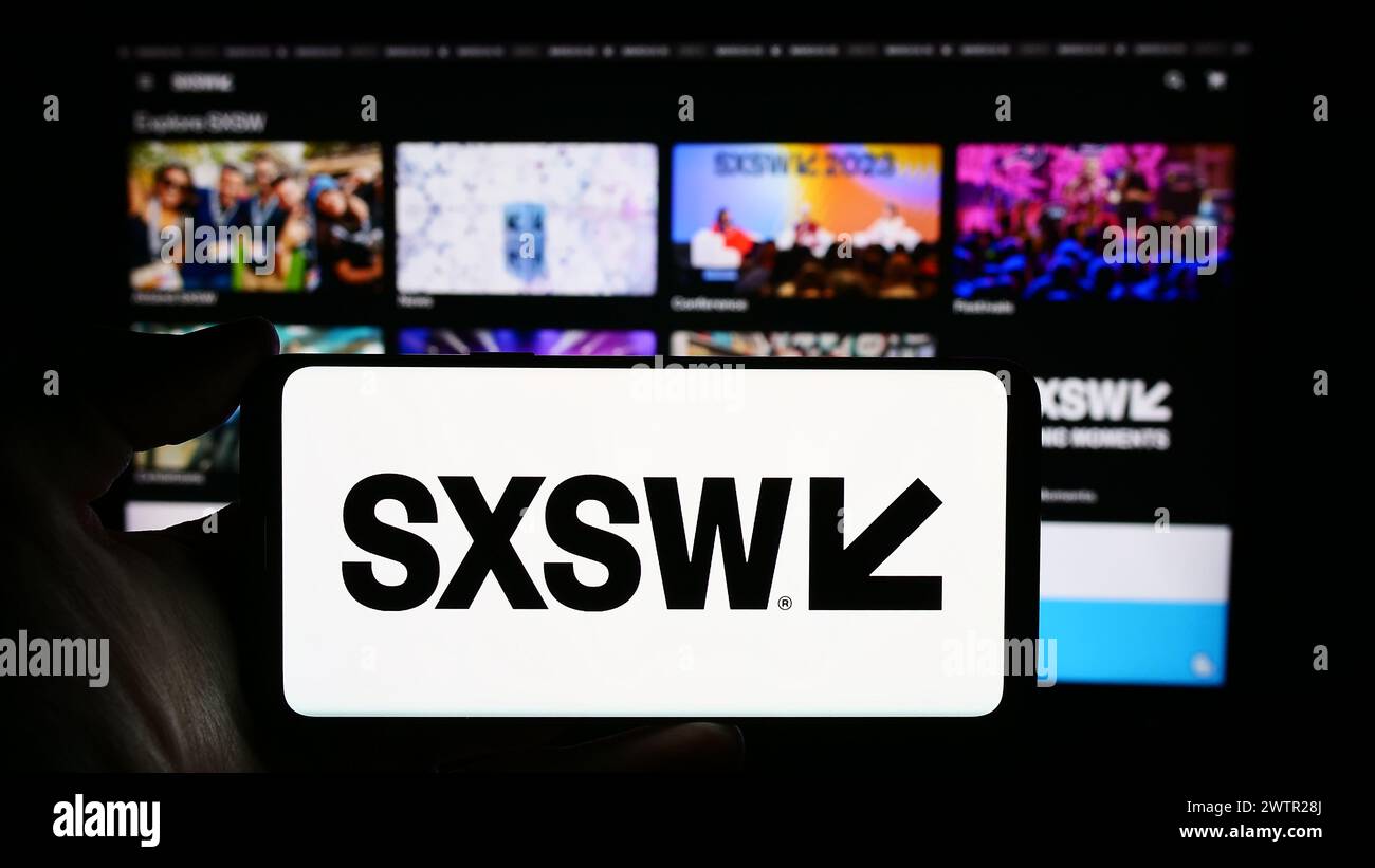 Person holding cellphone with logo of annual film and media event South by Southwest (SXSW) in front of webpage. Focus on phone display. Stock Photo
