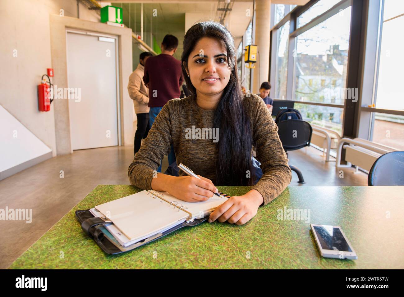 Young student making notes Aachen, Germany. Young, female Indian university exchange student working on a dormatory work desk, using her smartphone and analog notebook. MRYES Aachen Universiteit vsan Aachen Nortrhein-Westfalen Duitsland Copyright: xGuidoxKoppesx Stock Photo