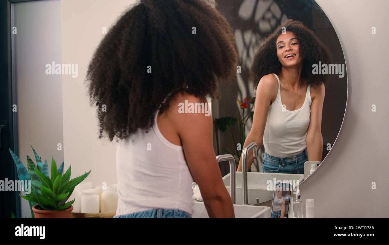 African American beautiful happy smiling pretty woman girl female with curly hair pampering looking at mirror morning beauty procedure record video Stock Photo