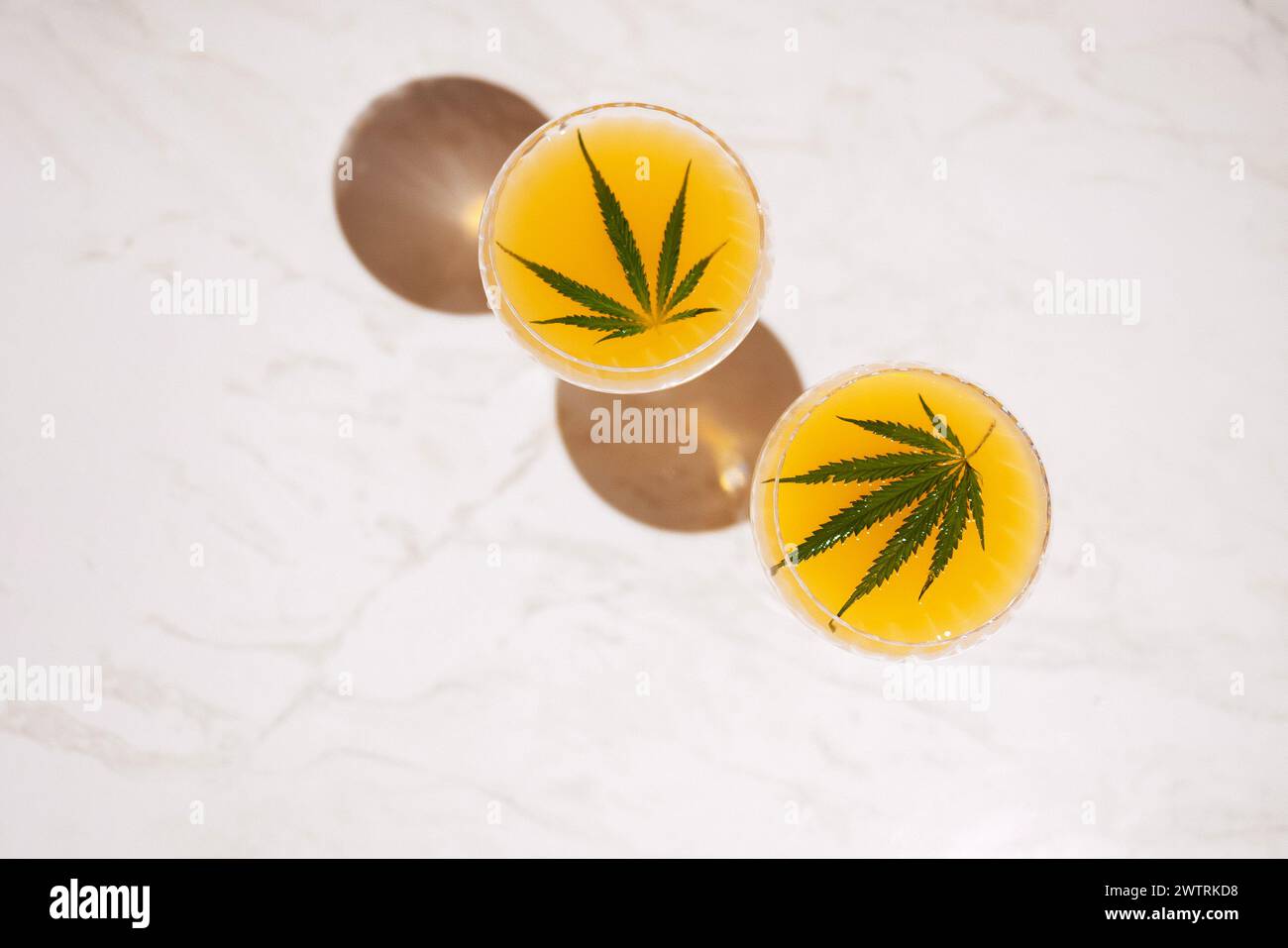 Two elegant long-stemmed glasses with green cocktails with cannabis leaves on the light marble surface of the table. A delicious healthy drink with he Stock Photo