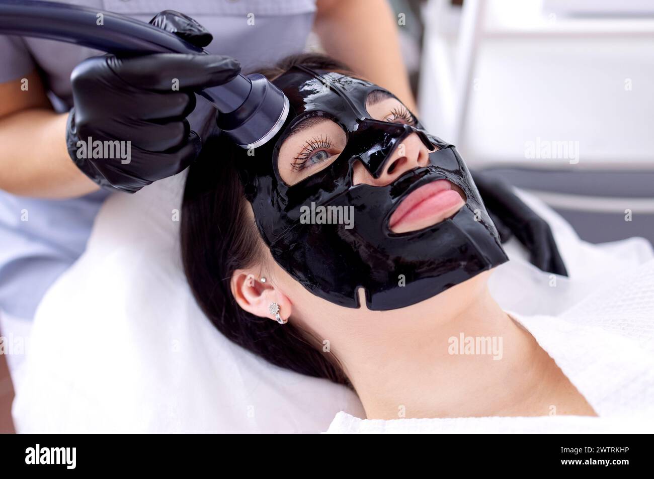 Close-up of a young woman in a black cosmetic mask at a spa procedure. A cosmetologist performs a cosmetic procedure using an ultrasound device. Moist Stock Photo