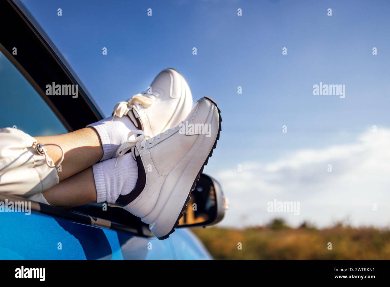 A close-up of a female legs in white sneakers are exposed to the car window. Traveling by automobile. Blue sky and a field in the background. Journey, Stock Photo