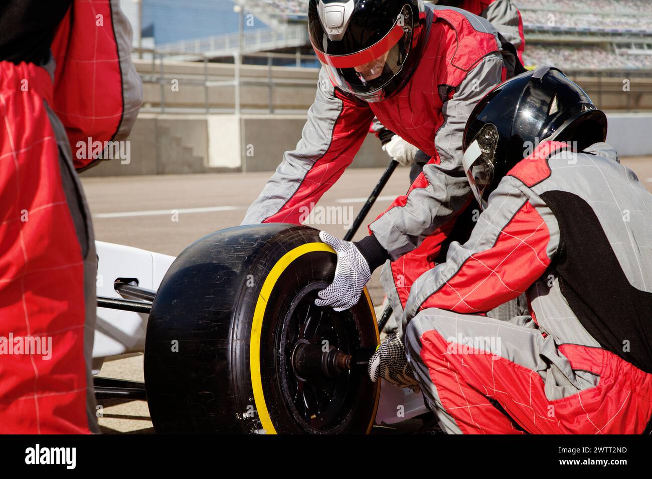 Pit crew members in red and white uniforms working on changing a tire of a white racing car on a racetrack. Stock Photo