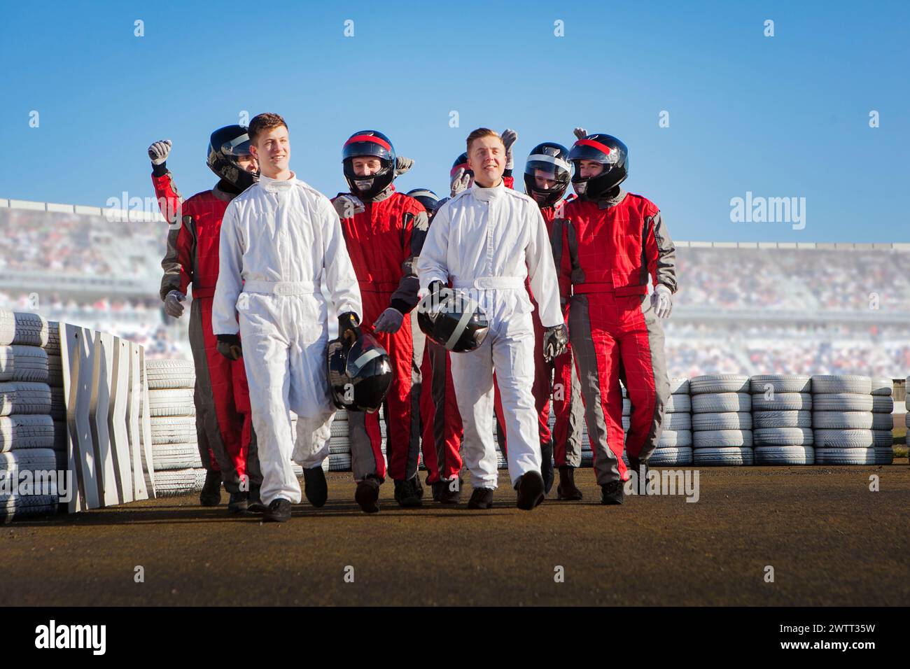 A group of people in racing suits walking on a racetrack, holding helmets and celebrating. Stock Photo