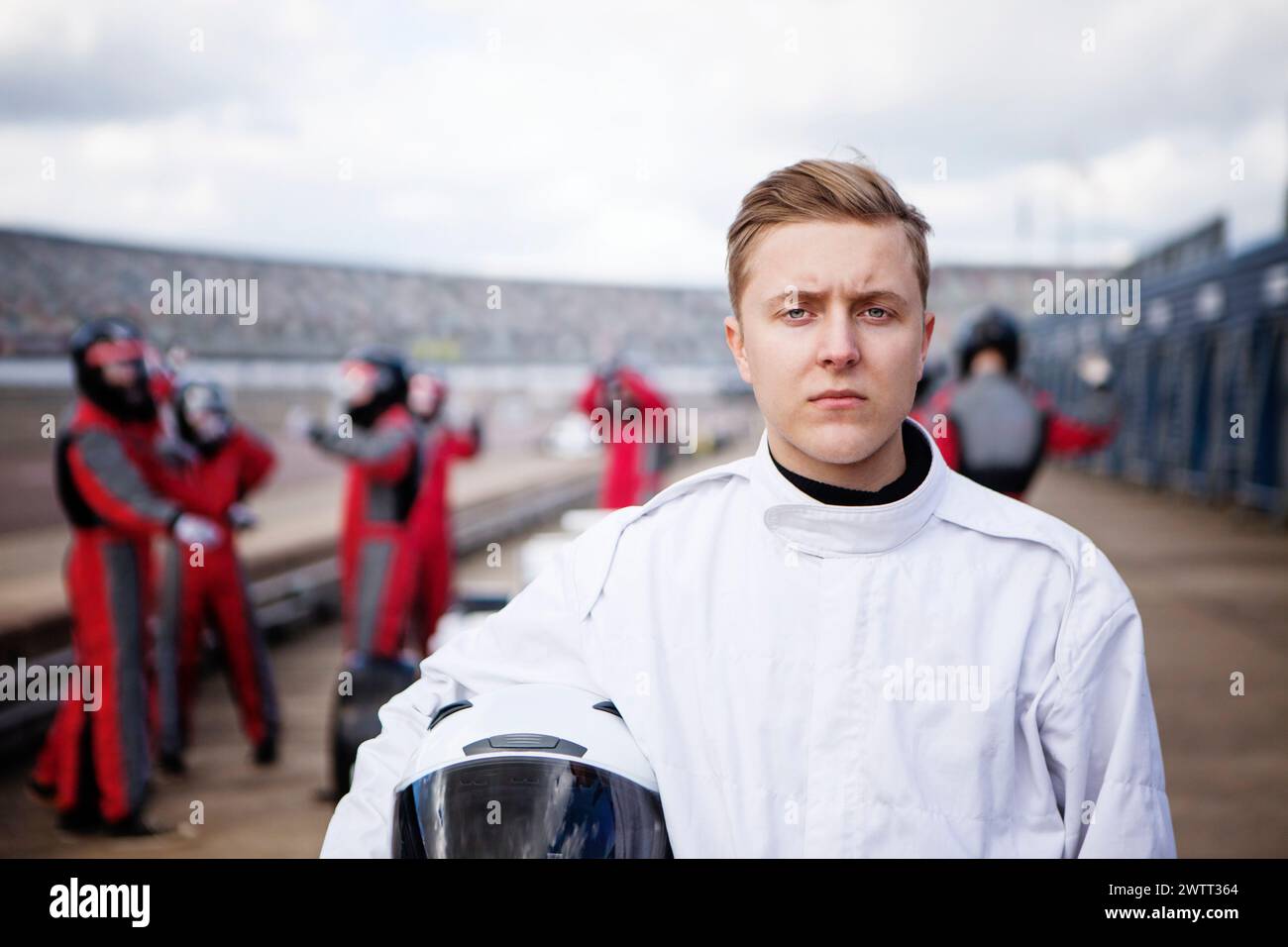Focused young racer holds helmet on racetrack with competitors in the background Stock Photo