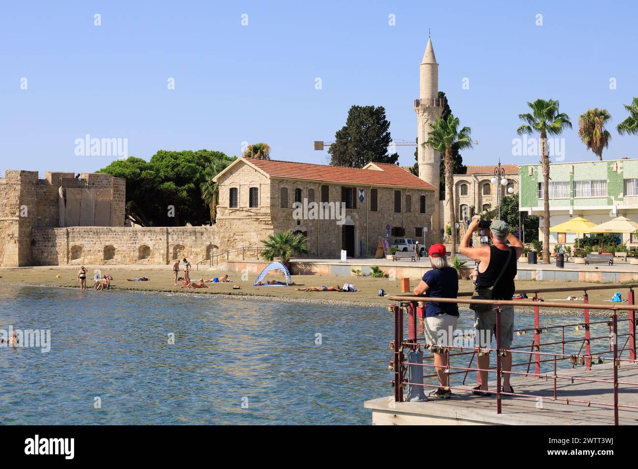 Tourist couple taking mobile phone picture of Larnaca Fort and the Mosque minaret. Sunbathers on the beach and swimmers. Finikoudes, Larnaca, Cyprus Stock Photo