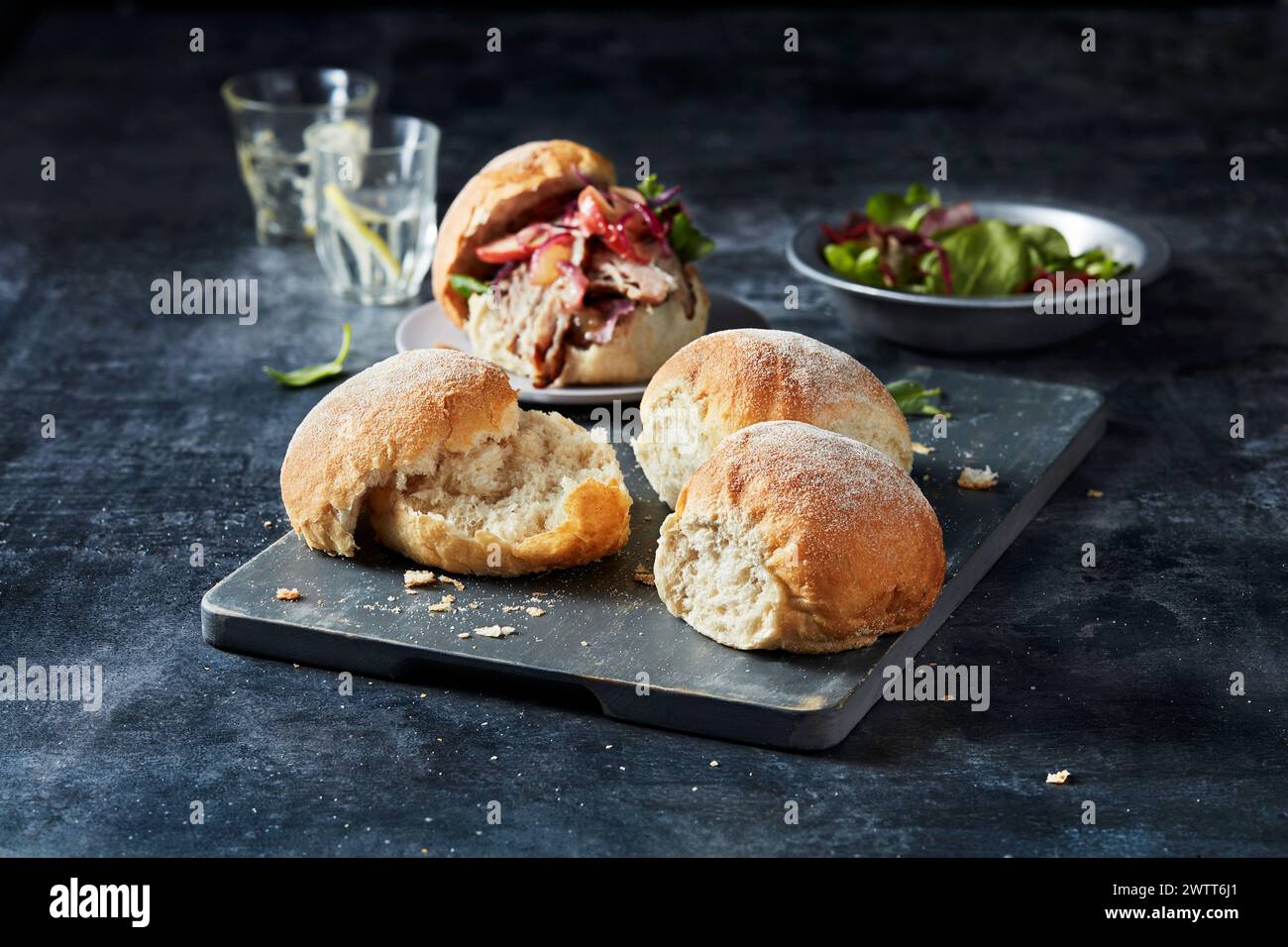 Gourmet pulled pork sandwiches on a rustic slate board with a side salad and a refreshing drink in the background. Stock Photo