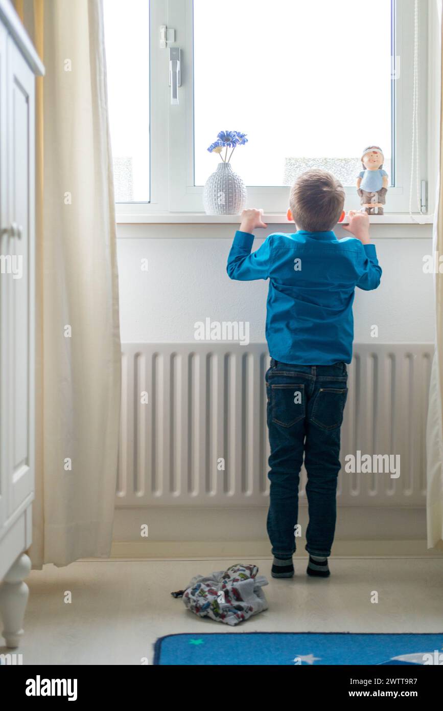 Young boy peering out of a bright window in a cozy room. Stock Photo