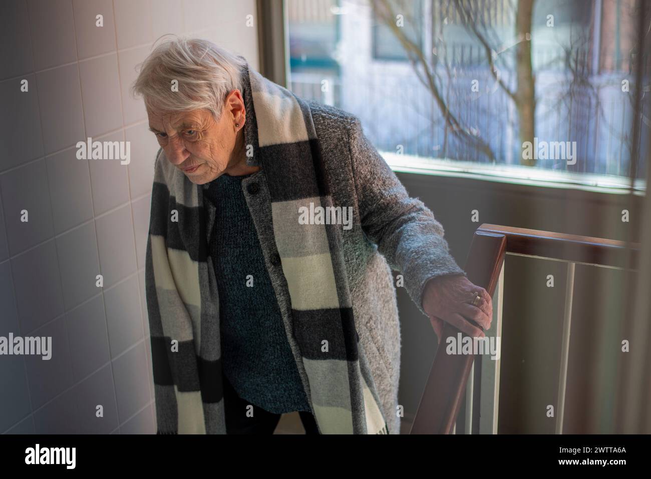 Elderly person standing by a staircase, bathed in soft natural light from a window. Stock Photo
