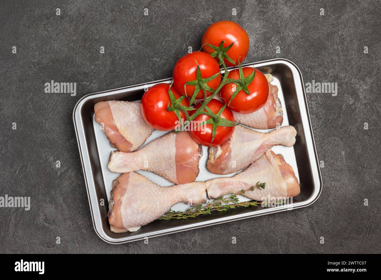 Raw chicken thighs in metal pallet. Green branch with tomatoes. Mushrooms in bowl. Black background. Flat lay Stock Photo