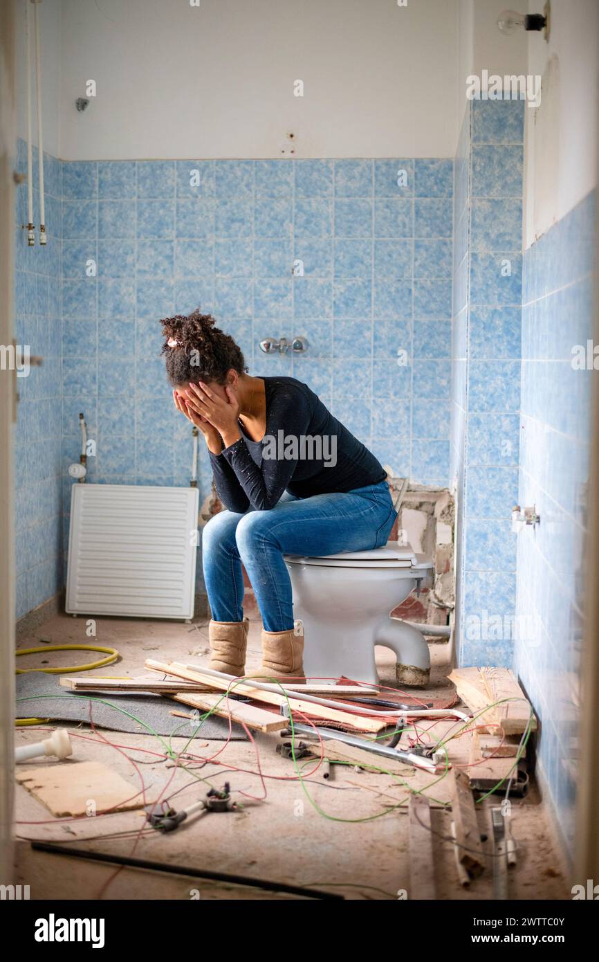 A person covers their face with their hands, seated on a toilet in a distressed state amidst a messy bathroom renovation. Stock Photo