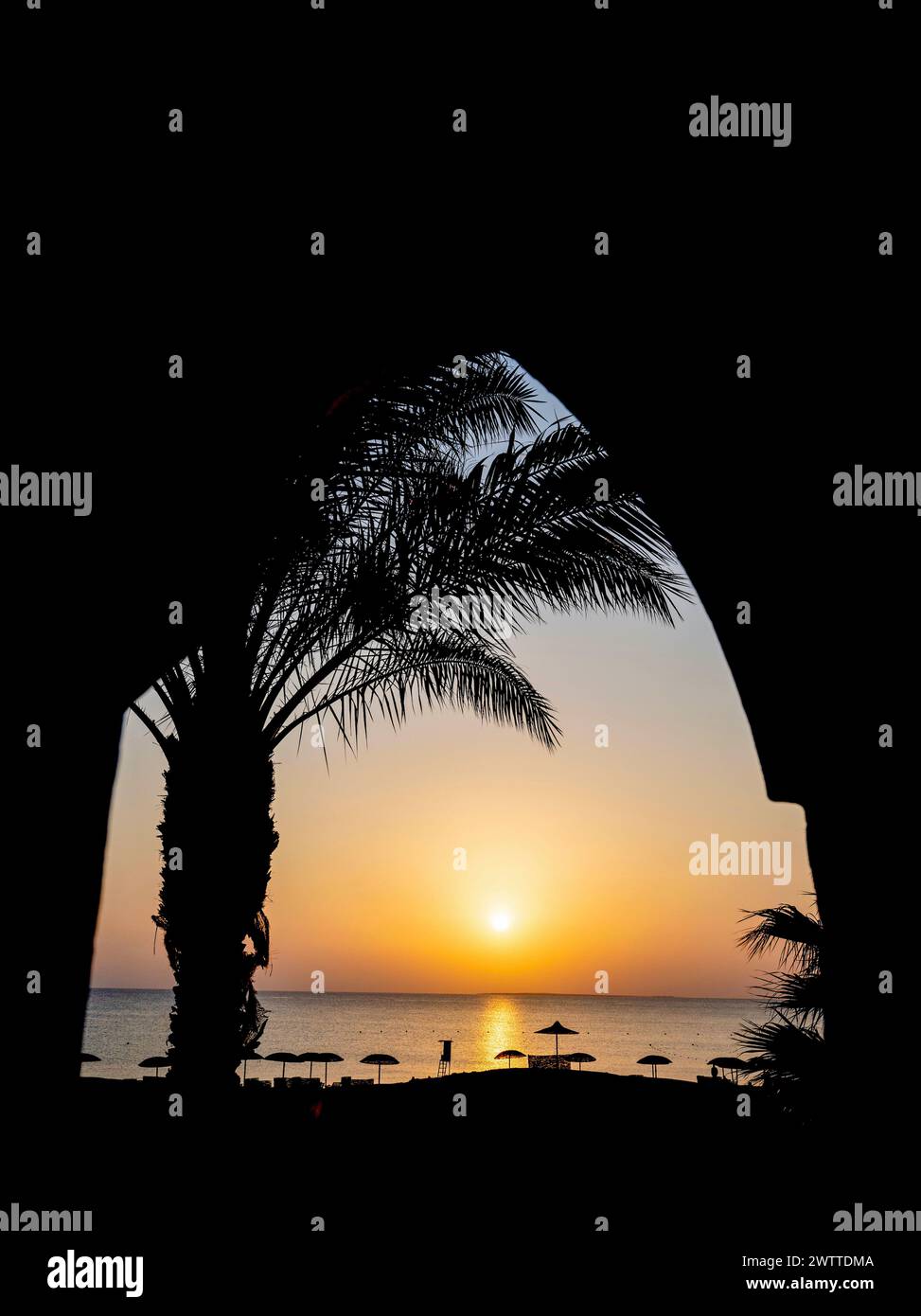 A serene sunset view through an archway framed by palm trees and beach umbrellas Stock Photo