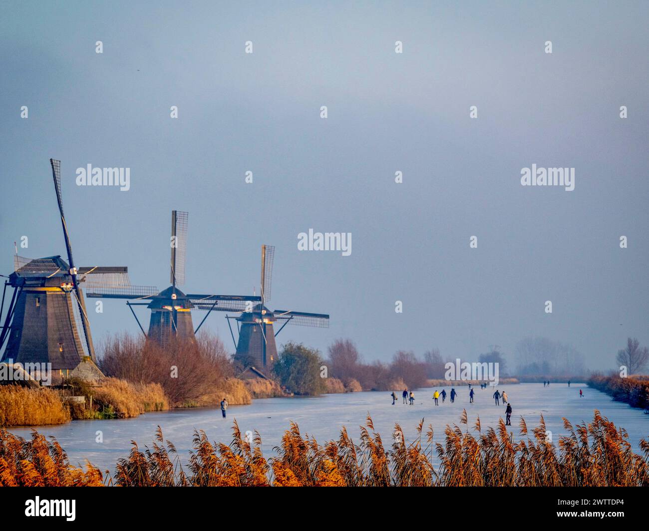 Skaters glide across a frozen canal with traditional windmills in the backdrop at dusk Stock Photo