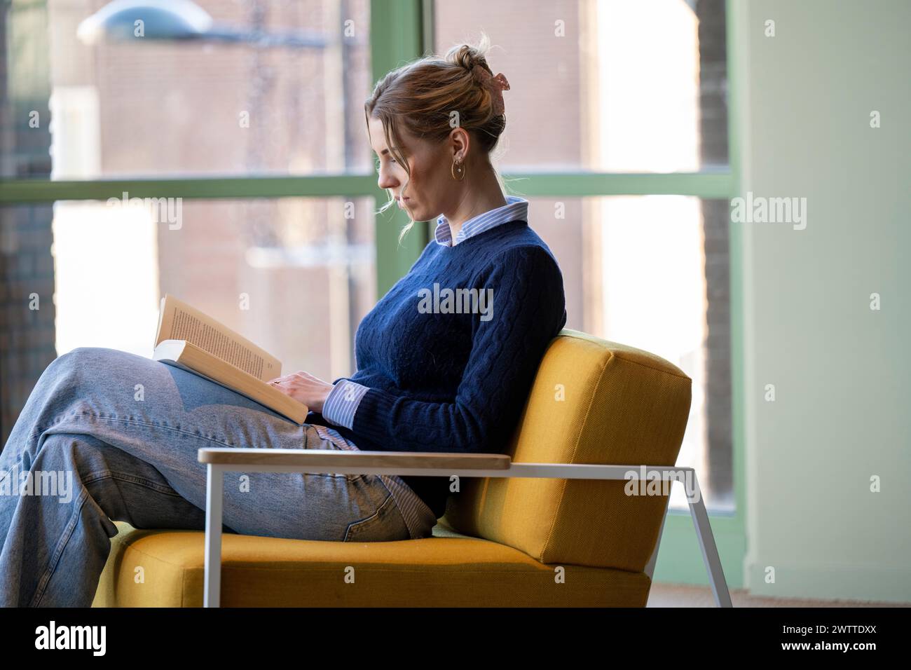 Person engrossed in a book while relaxing in a yellow chair. Stock Photo