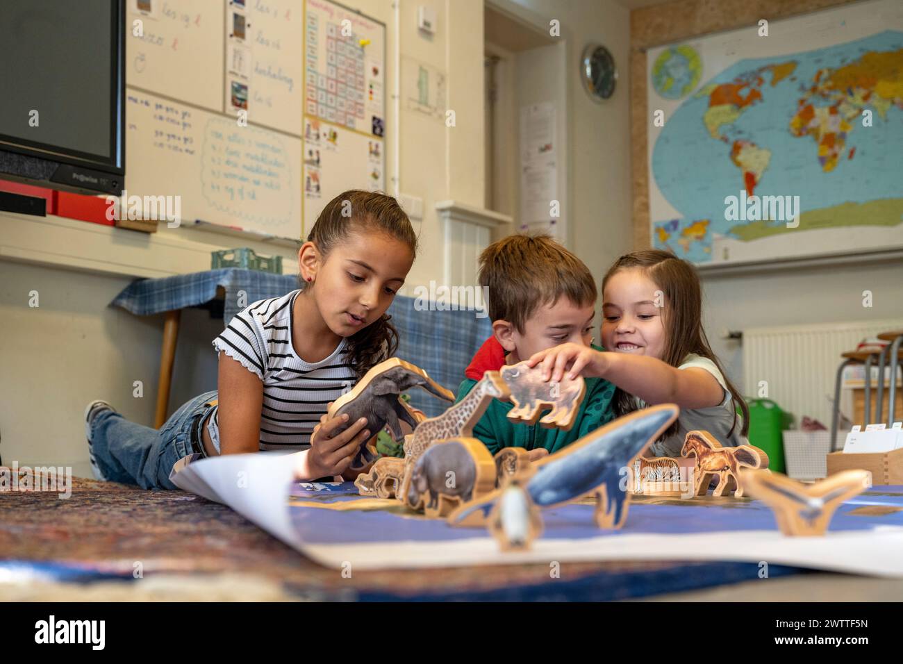 Three children are lying on the floor, engaging with various dinosaur toy models spread out on a carpet, with a world map in the background. Stock Photo