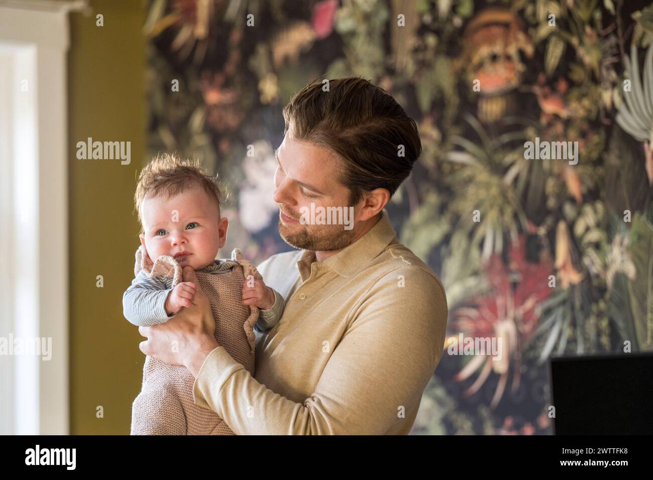 Young father holding his baby in a cozy room with a vibrant floral wallpaper background. Stock Photo