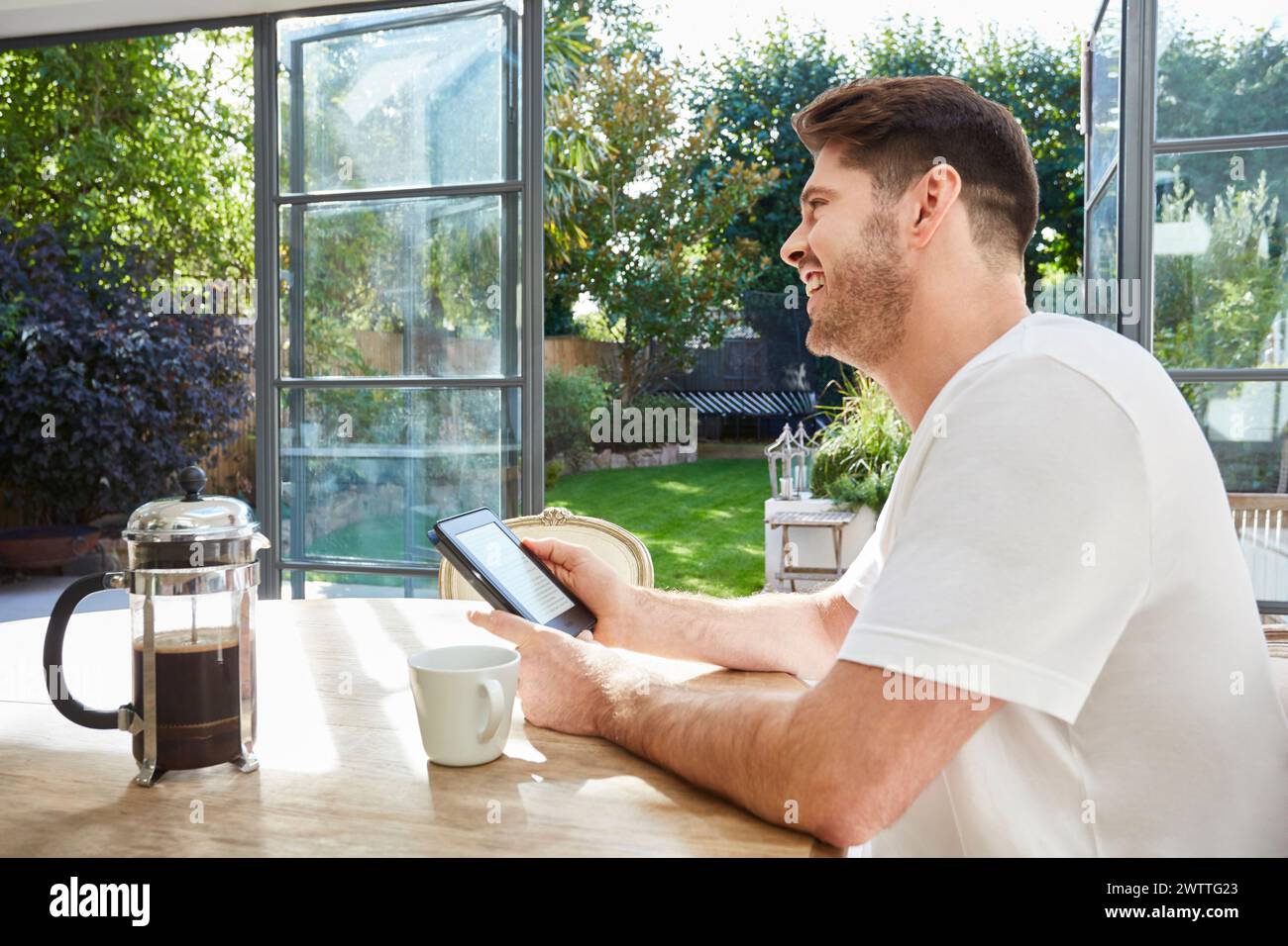 Man smiling while using a smartphone at a sunny kitchen table Stock Photo